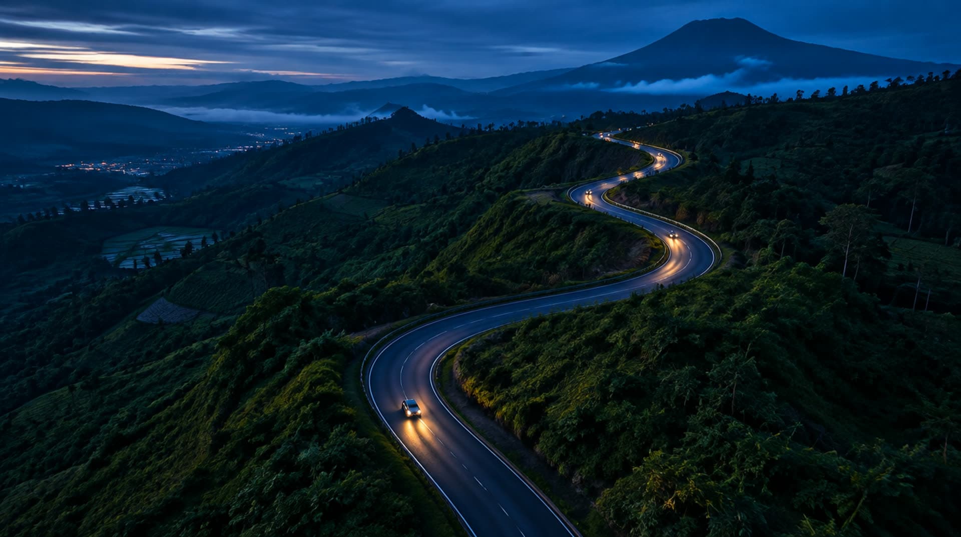 Jalan berkelok menuruni bukit hijau saat senja, lampu mobil menyala